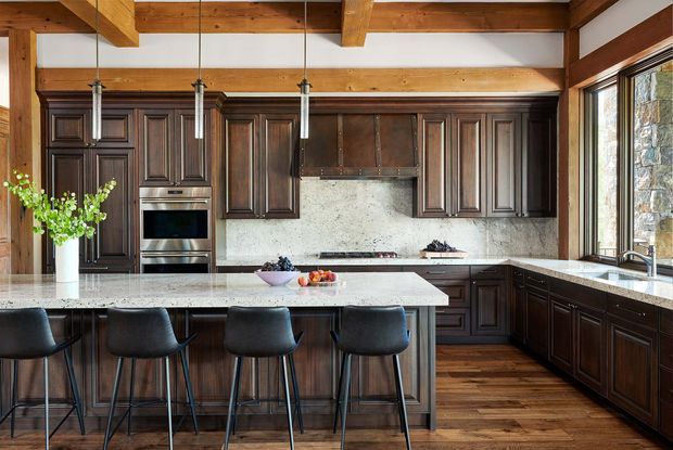 A spacious, richly detailed kitchen with dark raised-panel wood cabinetry, a large pale stone island with four bar stools, integrated stainless-steel appliances, a matching stone backsplash and benchtops, and exposed timber ceiling beams. Large windows bring in natural light, while pendant lights hang above the island. The room has a warm, refined, traditional lodge-inspired character.