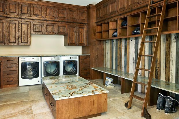 A rustic, richly timber-lined laundry room with extensive built-in cabinetry, three front-loading laundry appliances, a large central folding island with a decorative stone-like benchtop, open shelving, a long workbench, and a rolling library-style ladder. Vertical reclaimed timber boards form a feature wall, while large-format beige floor tiles complete the warm utility space.