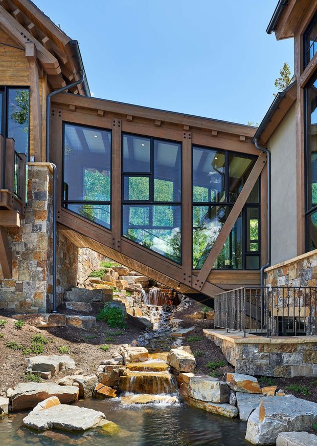 Exterior view of a multi-level timber-and-stone building complex connected by a glazed bridge spanning a landscaped rocky stream and waterfall. The bridge features dark timber structural framing, large black-framed windows, diagonal braces, and exposed connector plates. Stone-clad lower walls, timber eaves, and a small metal-railed terrace are visible under a clear blue sky.