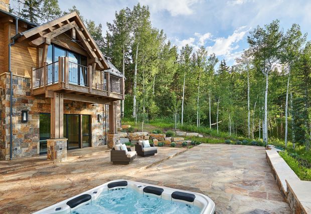 Exterior view of a rustic mountain-style residence surrounded by birch woodland. The two-storey facade combines natural stone masonry and warm timber cladding, with a prominent gabled roof, exposed rafters, timber balcony and expansive glazing. A broad stone-paved terrace includes outdoor seating and a partially visible spa pool, with landscaped retaining elements along the forest edge.