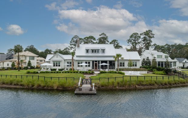 Wide exterior view of a large white residential property beside a calm waterway, with a central gable-fronted main volume, connected wings, white metal pitched roofs, dormer windows, extensive glazing, landscaped lawn, black boundary fencing, and a small waterfront landing. The composition has a polished coastal estate character under a bright blue sky.