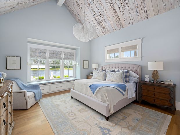 A bright, refined bedroom with a large upholstered bed, layered blue-and-white bedding, matching armchairs, bedside tables, and a broad patterned rug. The room features pale blue-grey walls, light timber flooring, built-in window seating and cabinetry beneath the windows, woven Roman blinds, and a distinctive whitewashed timber-clad vaulted ceiling with a textured pendant light.