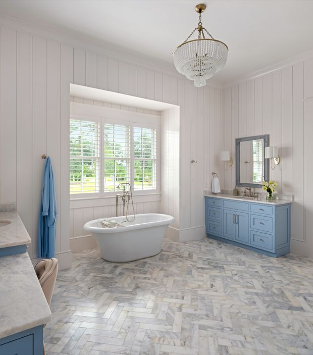 A bright, spacious bathroom with white vertical wall panelling, a freestanding white bathtub beneath plantation-shuttered windows, and a blue painted vanity with marble countertop. The floor is laid in a pale marble herringbone pattern, complemented by a framed mirror, wall sconces, and an ornate crystal chandelier. The overall look is refined, traditional, and light-filled.