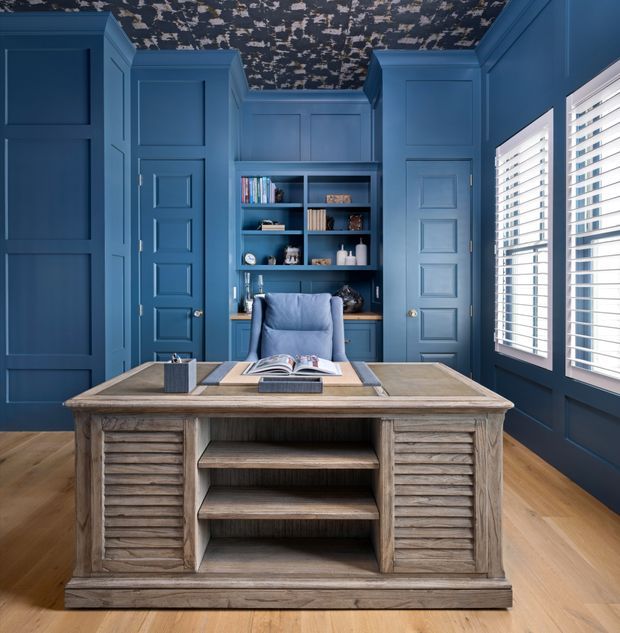 A richly styled blue home office with floor-to-ceiling panelled walls, built-in shelving, a central desk, and a blue upholstered chair. The room features warm timber flooring, a weathered timber desk with louvered drawer fronts and open shelving, white plantation shutters, and a dramatic dark patterned ceiling.