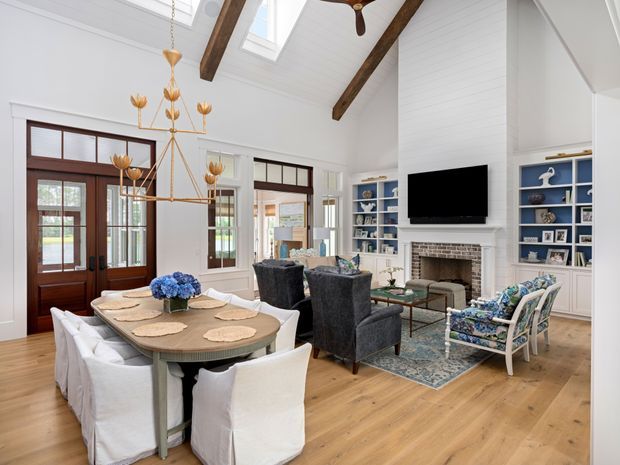 Bright open-plan dining and living room with a coastal-traditional feel. The space features a large oval timber dining table with slipcovered chairs, white paneled walls and ceiling, exposed dark timber beams, a skylight, timber-framed glazed doors, built-in white cabinetry with blue backing, a central fireplace with brick surround, television, patterned area rug, and a blue-and-white upholstered armchair. High pitched ceiling and abundant glazing create an airy, inviting atmosphere.