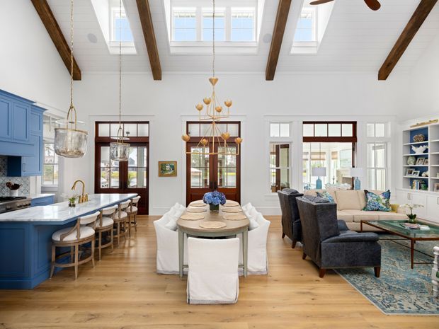 Bright open-plan interior combining a blue kitchen, dining area, and living room. The space has white walls and ceiling boards, warm timber flooring, exposed dark timber beams, abundant windows and skylights, white slipcovered dining chairs, dark lounge chairs, built-in shelving, and layered pendant lighting.