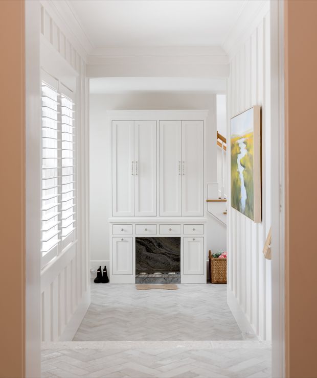 Bright, refined residential hallway or entry passage viewed through a framed opening. The space features white vertical wall panelling, detailed traditional mouldings, plantation shutters, a built-in white storage and display cabinet with a dark natural-stone fireplace or inset feature, and pale herringbone flooring. A colourful artwork and woven basket add restrained decorative accents. The corridor opens toward a stair or adjoining area in the background.