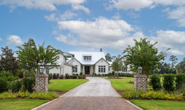 A large white country-style house viewed from the front, approached by a curved gravel driveway transitioning to a brick-paved entrance. The home has multiple gabled roof sections, white horizontal cladding, dormer windows, dark-framed multi-pane windows, and landscaped lawns with brick pillars and mature planting. Bright daytime exterior photography with a traditional, welcoming character.