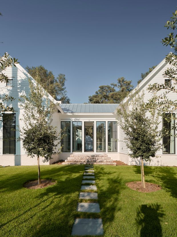 Front exterior view of a symmetrical white weatherboard residence arranged around a central glazed entrance pavilion. The building has steep gable roofs, a low metal-roofed connector, tall black-framed windows, brick entry steps, and a manicured lawn with a straight stepping-stone path and young trees. Clear daylight and mature vegetation create a refined, welcoming garden approach.