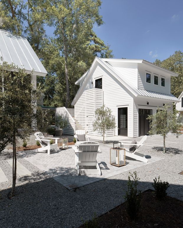 A bright landscaped courtyard with white gravel, pale paver paths, olive-like trees and white outdoor lounge chairs surrounding a modern white-clad house. The building features horizontal weatherboard siding, pitched gable forms, standing-seam metal roofing, dark-framed windows and doors, and a small covered entry. A second white-roofed structure is partially visible on the left.