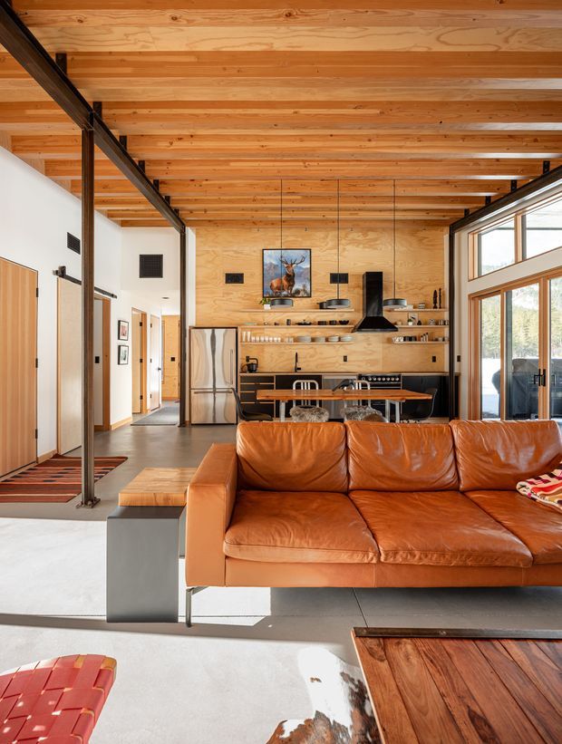 A bright open-plan living room with a visible kitchen and dining area. The space features extensive warm timber ceiling and wall finishes, exposed dark steel structural posts and beams, large glazed doors and windows, polished concrete flooring, and a prominent cognac leather sofa. The kitchen has timber cabinetry and open shelving, a dark rangehood, and stainless-steel appliances. The interior has a refined mid-century-influenced contemporary character with strong natural materials and indoor-outdoor glazing.