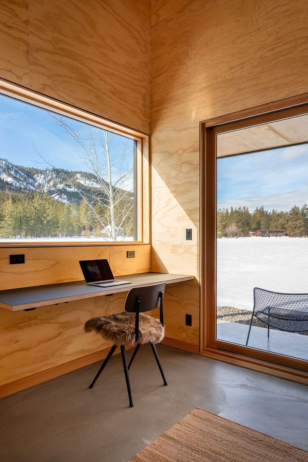 A compact, timber-lined workspace with a built-in wall desk, laptop, and black chair positioned beside large glazed openings. The room has warm plywood walls and window/door frames, a smooth grey concrete floor, and expansive views of a snowy mountain and forest landscape. A glazed door opens to an outdoor terrace with a second chair.