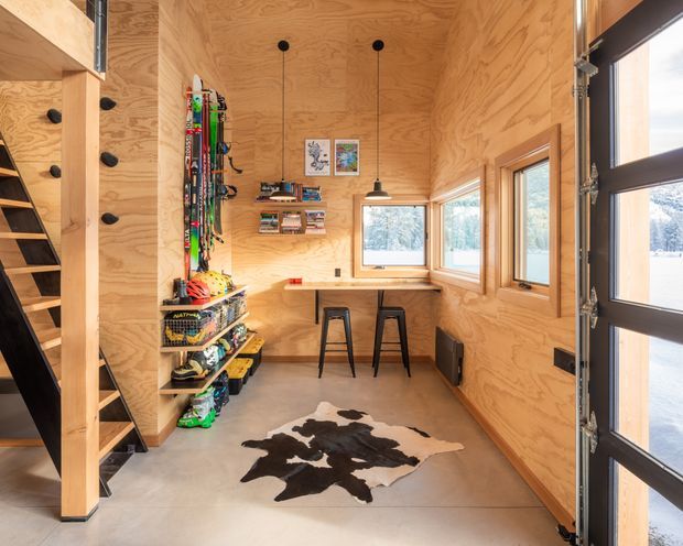 Compact wood-lined entry and storage space with a built-in open shelving unit, staircase, sports equipment storage, wall-mounted shelves, a small desk with two stools, multiple windows, and a glazed exterior door. The room has plywood wall and ceiling finishes, a smooth concrete floor, black pendant lights, and a cowhide rug.