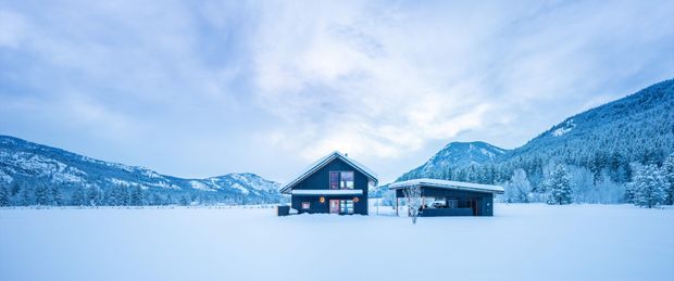 Wide exterior view of a dark timber-clad alpine house and attached low-profile carport or garage, set in a snow-covered open landscape with forested mountains behind. The building has a simple pitched gable roof, contrasting light trim, and large glazed openings. The scene is cool-toned, serene, and minimal, with the architecture clearly presented against the expansive snowy setting.