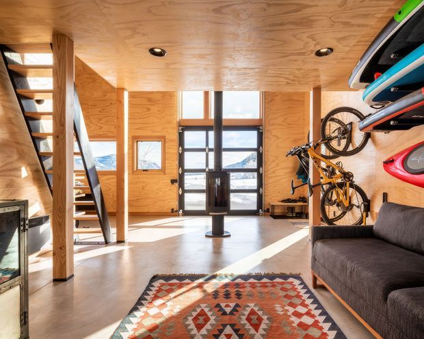 A bright, compact multi-purpose entry and gear-storage space with plywood-lined walls and ceiling, polished concrete flooring, a sculptural open timber stair, large glazed entry doors, wall-mounted bicycles and surfboards, and a dark upholstered sofa. Mountain scenery is visible through the glazing. A patterned rug adds warmth to the otherwise practical, contemporary interior.