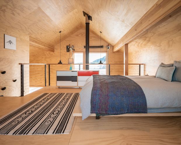 A compact loft-style bedroom lined extensively in warm plywood, with a low platform bed in the foreground, patterned monochrome rug, built-in storage, climbing holds on the wall, a central black wood-burning stove flue, pendant lights, and a timber-framed mezzanine railing. A small window provides a view beyond.