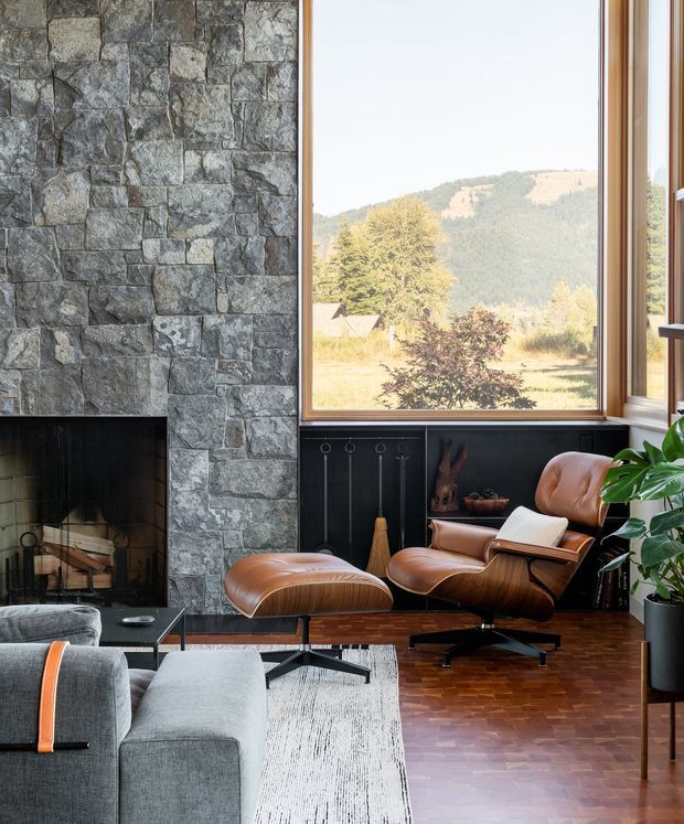 A refined living room with a dramatic floor-to-ceiling rough stone fireplace wall, expansive timber-framed picture windows, dark built-in cabinetry, and a mid-century leather lounge chair with matching ottoman. The room overlooks a wooded, mountainous landscape and features warm parquet timber flooring, a pale textured rug, a grey sectional sofa, and indoor greenery. The composition feels warm, sophisticated, and strongly connected to the surrounding natural setting.