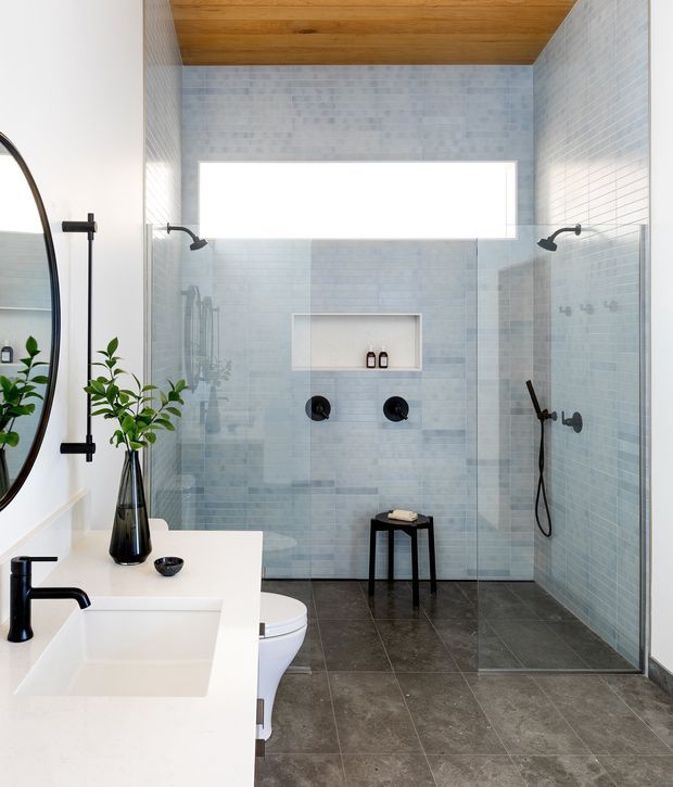 Bright contemporary bathroom with a large glass-enclosed walk-in shower, pale blue-grey tiled walls, dark grey tiled floor, white floating vanity with integrated rectangular basin, matte black tapware and shower fittings, and a warm timber-lined ceiling. A round mirror, black stool, plant, and recessed shower niche complete the clean modern scheme.