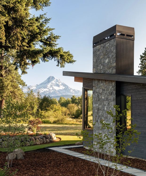 Exterior view of a contemporary mountain house with a tall stone-clad chimney or tower, dark horizontal timber cladding, broad flat roof overhangs, and large glazed openings. The building is set within a landscaped garden, with a prominent snow-capped mountain visible in the background.