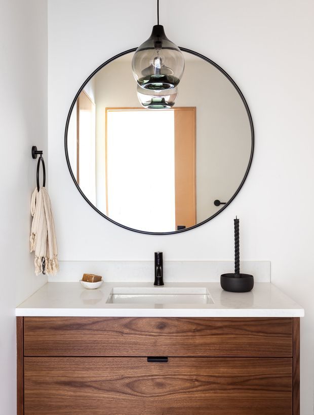 A refined bathroom vanity vignette featuring a floating dark timber drawer cabinet, white integrated rectangular basin and benchtop, matte black tapware, fittings and accessories, and a large round black-framed mirror. A sculptural glass pendant light hangs in front of the mirror, while a textile towel and minimal decorative objects add texture. The palette is predominantly white, warm timber brown and black, with a clean contemporary feel.