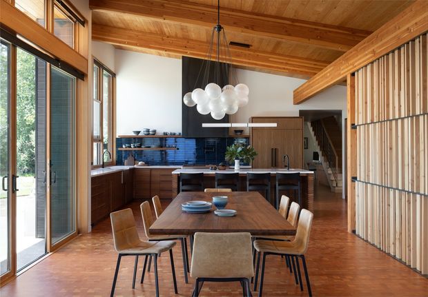 Open-plan dining and kitchen interior with a long timber dining table and mixed upholstered dining chairs in the foreground. The kitchen features dark wood cabinetry, a blue tile backsplash, a central island, and integrated appliances. Exposed timber ceiling beams, warm timber flooring, extensive glazing, and a sculptural cluster pendant light create a warm, refined mid-century-inspired atmosphere. A timber slatted screen and stairway are visible at the right.