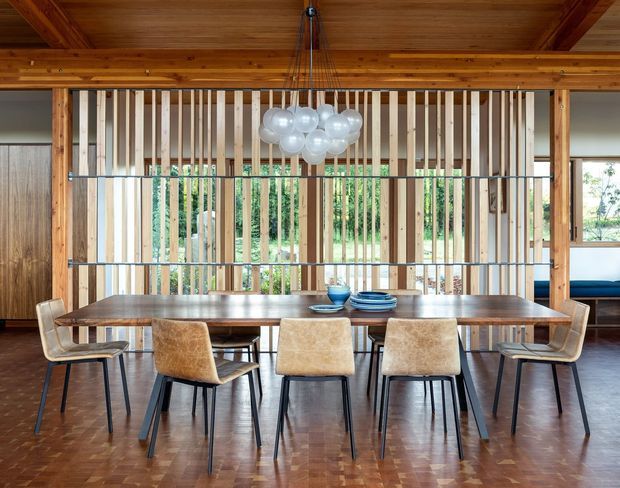 A warm, timber-lined dining room centred on a long dark wood dining table with six upholstered chairs. A sculptural cluster pendant hangs above the table, while a vertical timber slat screen separates the dining area from the adjoining space and allows views through to greenery outside. The room has a polished parquet timber floor, exposed timber ceiling beams, and a refined contemporary feel.
