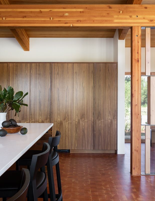 Interior view of a warm, timber-rich dining and kitchen area. A long white countertop with dark dining chairs occupies the foreground, while full-height dark timber cabinetry lines the back wall. Exposed timber beams, ceiling boards, and structural posts frame the space, with parquet timber flooring and tall glazed openings to the right.