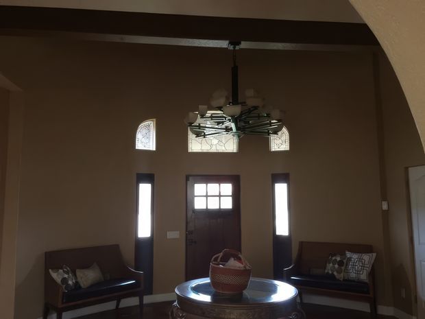 A dim, formal entry hall viewed from inside, with a dark wood front door flanked by narrow sidelights and topped by small decorative stained-glass windows. Two upholstered wooden benches sit against the walls, while a round central table with a decorative bowl anchors the space. A heavy exposed dark beam spans the ceiling, and an arched opening frames the room.