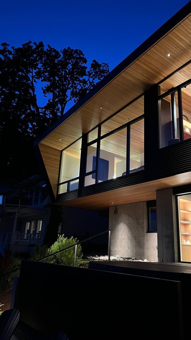 Nighttime exterior view of a contemporary two-storey house with a strongly cantilevered upper level, extensive black-framed glazing, warm timber-lined soffits and ceilings, and exposed concrete walls. Recessed lights illuminate the covered terrace and entry area, while mature trees and a neighbouring residence form the dark background.