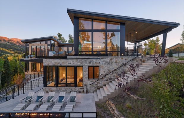 Exterior view of a contemporary multi-level hillside residence with expansive floor-to-ceiling glazing, dark metal roof and frames, extensive natural stone walls, broad terraces, exterior stairs, and a poolside deck with lounge chairs. The house is set on a steep, landscaped site with mountain terrain visible in the background.