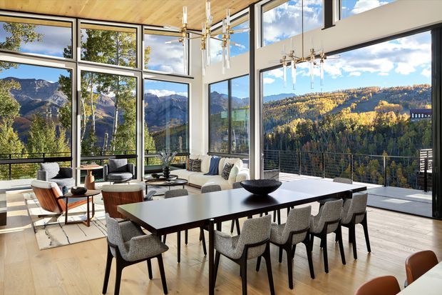 A bright, sophisticated open-plan living and dining space with expansive floor-to-ceiling glazing overlooking a dramatic mountainous, forested landscape. The room features a large dark dining table with upholstered dining chairs, a neutral sectional sofa, mid-century-inspired lounge chairs, light timber flooring, a timber-lined ceiling, and sculptural contemporary pendant lighting. A balcony or terrace with dark metal balustrading is visible outside.