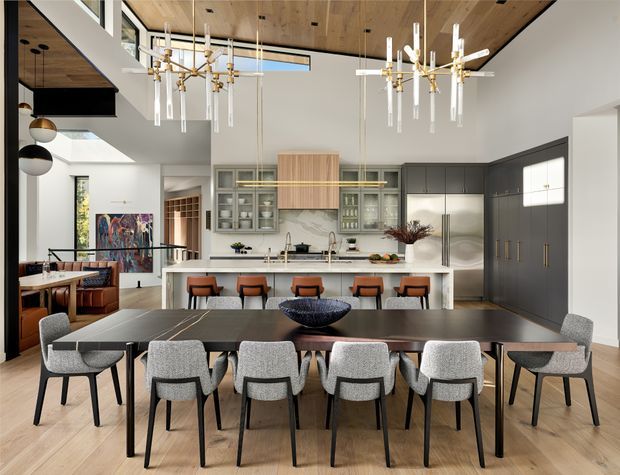 Spacious open-plan dining and kitchen interior with a long dark dining table and upholstered dining chairs in the foreground, facing a large kitchen island with bar seating. The room has a high ceiling with exposed timber lining, dramatic contemporary pendant lights, dark built-in cabinetry, a pale island, extensive glazing, and an adjoining lounge area visible to the left. The design combines refined contemporary detailing with warm timber surfaces and muted neutral furnishings.