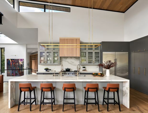 A spacious contemporary kitchen with a large marble island and five orange upholstered bar stools. Dark grey floor-to-ceiling cabinetry, integrated stainless-steel appliances, glass-front display cabinets, and a marble splashback frame the cooking zone. Long linear pendant lights hang over the island, while a warm timber ceiling and timber flooring add contrast. The open-plan space has high ceilings and clerestory glazing, with artwork and adjacent living areas visible in the background.