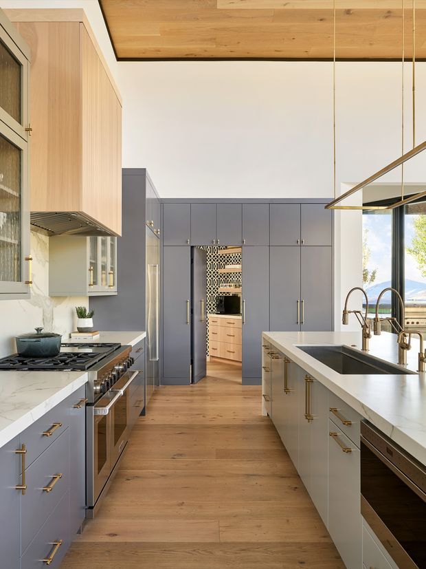 A refined contemporary kitchen with a long central island, integrated double sink and brass-toned tapware, extensive grey-blue cabinetry, and a second run of cabinetry along the left wall. Warm timber flooring and a timber-lined ceiling contrast with pale stone benchtops, while a large window brings in natural light and exterior views. A patterned opening connects to a pantry or secondary preparation area at the rear.