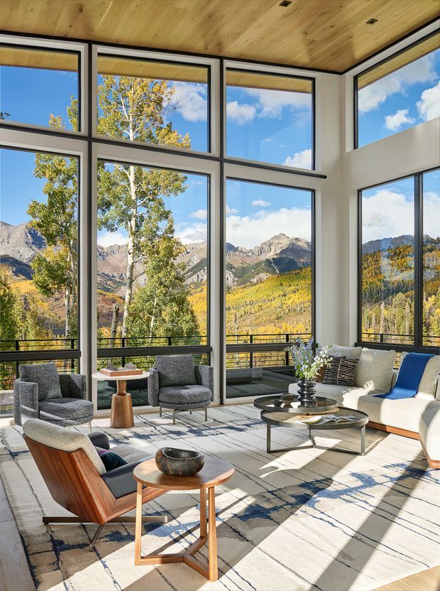 Bright contemporary living room with expansive floor-to-ceiling corner glazing overlooking autumnal mountain scenery. The room has a warm timber-lined ceiling, pale walls, dark window frames, a large light rug with geometric navy patterning, and a refined arrangement of upholstered lounge chairs, a sofa, and low round coffee tables.