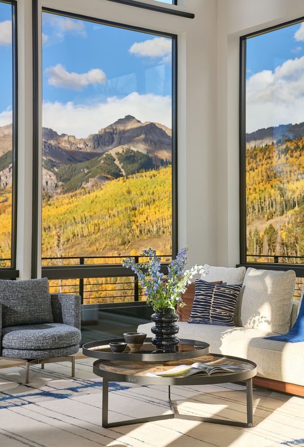 A bright, contemporary living room with expansive black-framed floor-to-ceiling windows overlooking an autumn mountain landscape. The room contains a light upholstered sofa, grey lounge chair, nested round coffee tables, patterned cushions, a large pale rug, and a floral arrangement. Strong natural light and the panoramic mountain view create an airy, refined atmosphere.