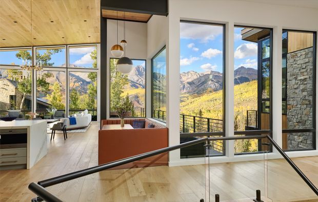 A bright contemporary open-plan interior with a kitchen at left, living area beyond, and a stair or landing in the foreground enclosed by glass balustrades and dark metal handrails. Floor-to-ceiling glazing frames a dramatic mountain landscape, while a warm timber ceiling, pale timber flooring, white walls, black-framed windows, pendant lights, and a stone-clad exterior wall create a refined modern alpine atmosphere.