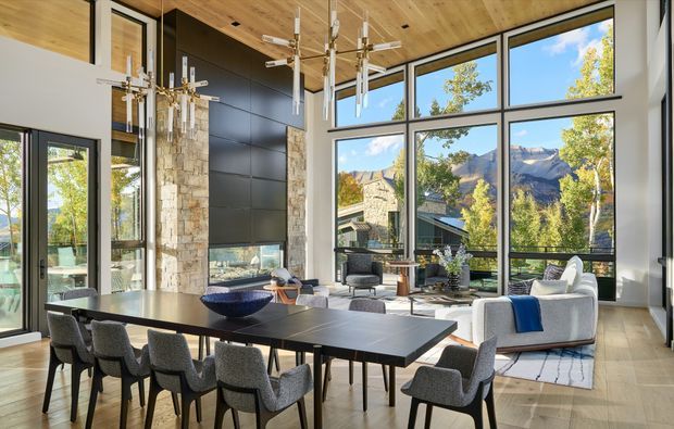 A spacious, light-filled open-plan living and dining room with dramatic floor-to-ceiling glazing overlooking a mountainous landscape. The room features a long dark dining table with upholstered chairs, a contemporary lounge area, a tall stone-clad fireplace wall, dark panelled joinery, a timber-lined ceiling, and sculptural pendant lights.
