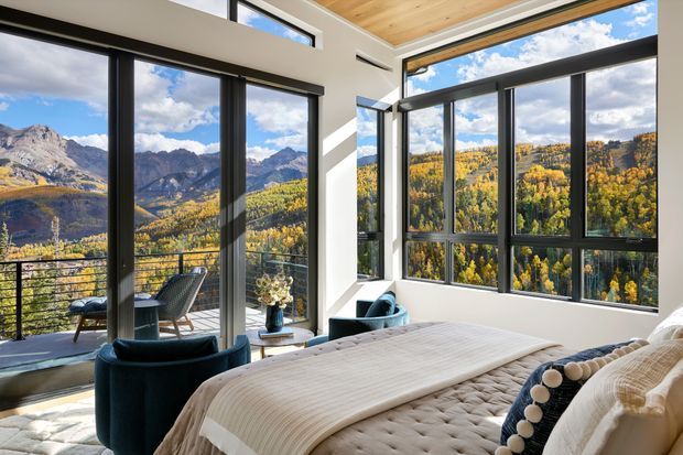 A bright, contemporary bedroom with floor-to-ceiling black-framed glazing overlooking an expansive alpine mountain and autumn forest landscape. The room features a large upholstered bed with layered neutral bedding, deep teal lounge chairs, a small side table, white walls, and a warm timber-lined ceiling. A balcony with outdoor seating is visible beyond the glazing.