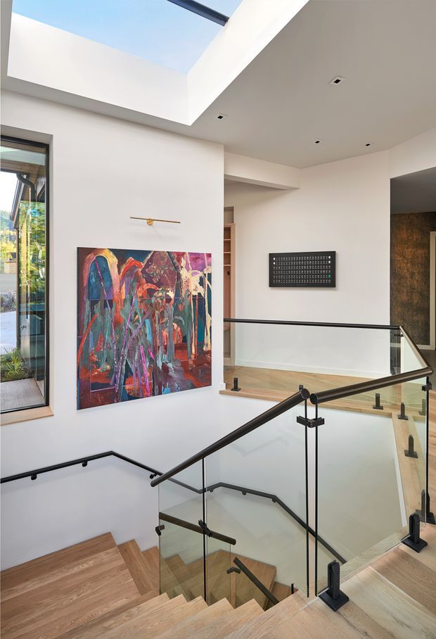 A bright, contemporary multi-level stair hall with an open stairwell, light timber stair treads and flooring, clear glass balustrades, and slim black metal handrails. White walls and ceilings surround a large colorful abstract artwork, while a dramatic skylight or roof opening brings in daylight. A narrow vertical window and adjacent shelving are visible, with a darker textured feature wall at the upper-right edge.
