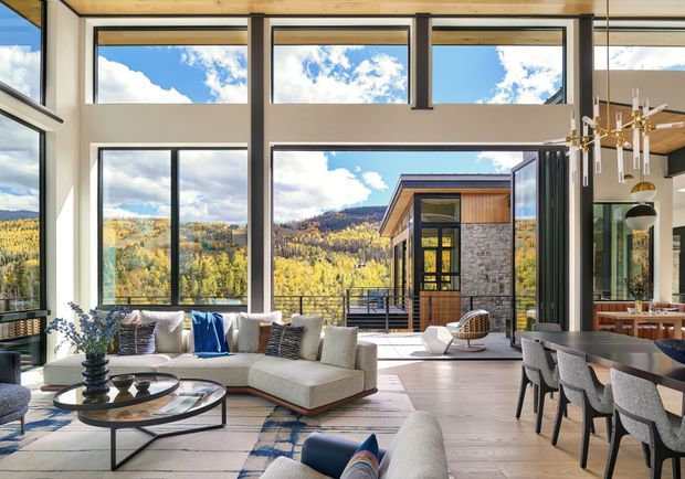 A bright, expansive open-plan living and dining room with floor-to-ceiling glazing and dramatic clerestory windows. A large pale sectional sofa, low round coffee table, contemporary dining table with upholstered chairs, and sculptural pendant lighting sit within a warm timber-framed interior. The glazing opens to a terrace and frames a mountainous, autumnal landscape. The room has a high ceiling and strong indoor-outdoor connection.