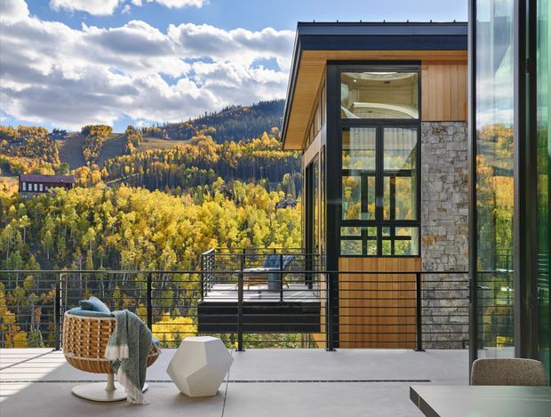 A contemporary mountain home interior opening onto a broad terrace, with floor-to-ceiling glazing, black-framed windows and doors, vertical timber cladding, and a natural stone wall. The terrace overlooks a forested mountain landscape in autumn. A sculptural white side table, woven lounge chair, throw, and part of a dining table are visible in the foreground.