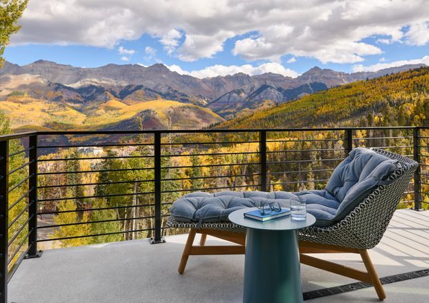 A modern outdoor terrace or balcony with a black horizontal-bar metal balustrade, pale concrete flooring, and a contemporary cushioned lounge chair with ottoman beside a small round side table. The elevated setting overlooks a dramatic mountain valley covered in autumn foliage under a partly cloudy blue sky.