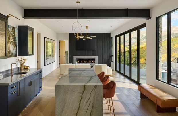 Open-plan contemporary kitchen and living area with a large stone island, dark blue fixed cabinetry, timber flooring, black steel-framed glazing, and a dark ribbed feature wall with a linear fireplace. Warm leather bar stools and a bench seat contrast with the neutral finishes, while expansive glazing provides views of a wooded mountain landscape.
