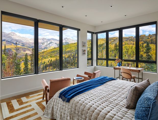 A bright contemporary bedroom with expansive black-framed corner windows overlooking a dramatic mountain and forested landscape. The room contains a large bed with a textured white quilt, blue throw and cushions, a pair of upholstered lounge chairs, a small round side table, and a compact timber desk with a chair. Recessed ceiling lights, light neutral walls, timber flooring, and a geometric area rug create a clean, comfortable retreat.
