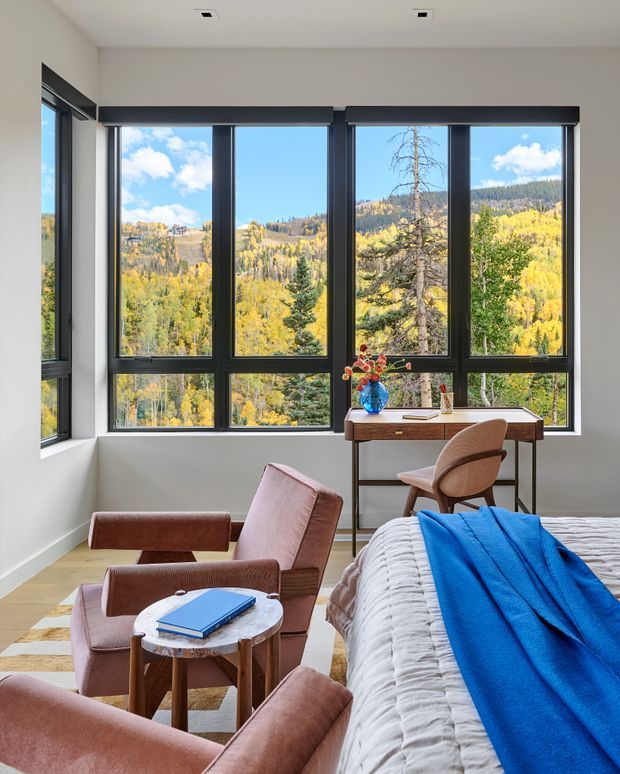 A bright, contemporary bedroom with expansive black-framed corner windows overlooking a wooded mountain landscape in autumn. The room includes a bed with a textured white cover and vivid blue throw, a compact wooden writing desk with a curved upholstered chair, and two rust-toned lounge chairs around a small round marble-topped table. White walls, recessed ceiling lights, and warm timber flooring create a refined, airy retreat.
