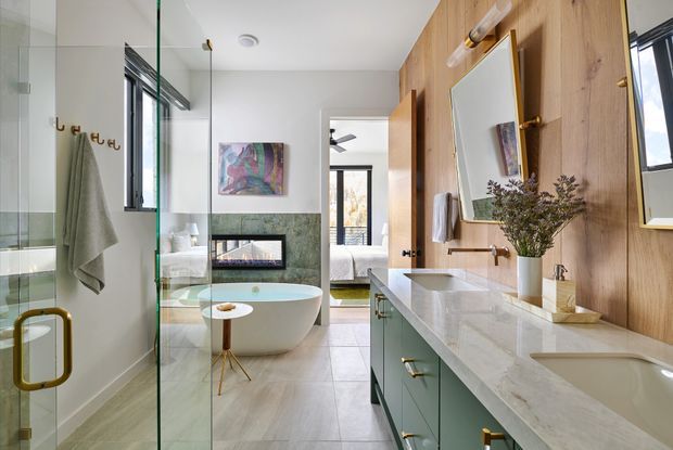 A bright contemporary bathroom viewed from the vanity area, featuring a glass-enclosed shower, freestanding oval bathtub, double vanity, pale stone benchtop, green cabinetry, and warm timber wall panelling. A bedroom is visible through the open doorway beyond the bathroom.