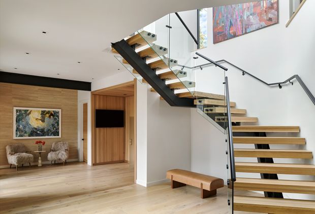 A bright contemporary open-plan interior featuring a sculptural floating timber staircase with black steel stringers, clear glass balustrades, and minimalist black handrails. Pale timber flooring continues through the space, with a timber-lined built-in media cabinet and warm timber feature wall. Artwork, upholstered lounge chairs, and a bench add visual interest beneath a high ceiling.
