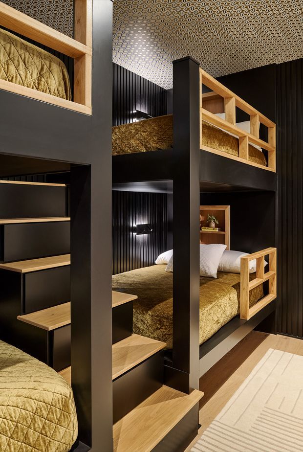 A compact multi-level bunk bedroom with four built-in sleeping platforms arranged around a central stair and storage structure. The room features black vertical wall panelling, matte black structural framing, warm timber stair treads and bunk rails, patterned geometric ceiling wallpaper, and quilted olive-gold bedding. Integrated wall lighting and open shelving create a refined, dramatic lodge-like interior.