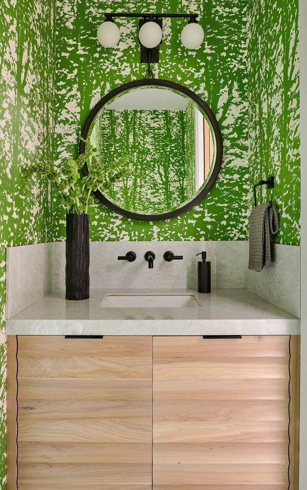 A compact, boldly styled bathroom vanity with a pale stone benchtop and integrated white basin, light timber drawer cabinetry, matte black wall-mounted tapware, and a large round black-framed mirror. The walls feature a vivid distressed green-and-white patterned finish, complemented by a black three-globe wall light, dark sculptural vase, and hanging towel.